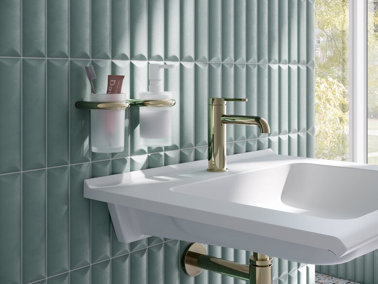 Wandhalterungen mit Becher und Seifenspender neben Waschbecken, Wasserhahn und Siphon in messingfarbener Optik an blauer Fliesenwand