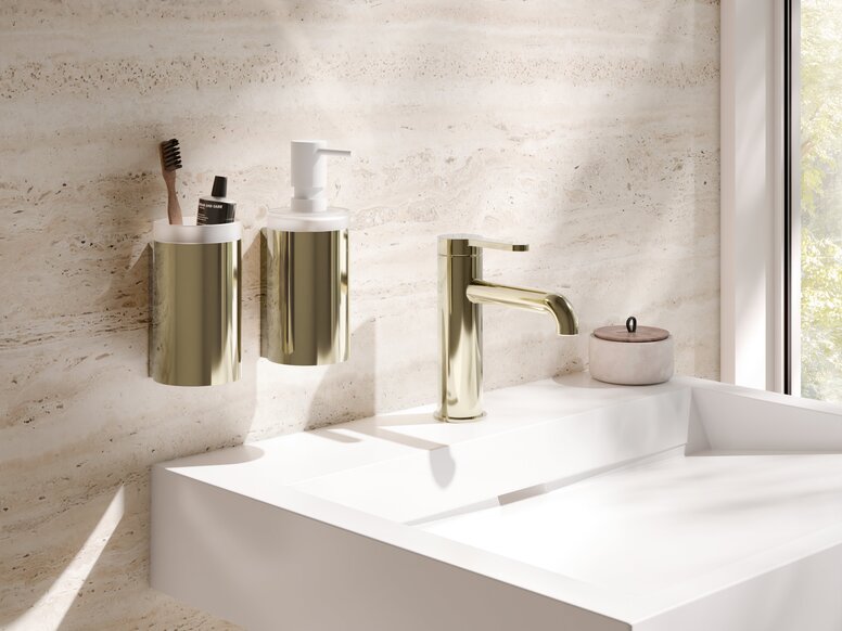 HEWI PVD AQ477 fittings A brass-coloured PVD toothbrush holder and soap dispenser next to a tap on a white washbasin against a beige wall