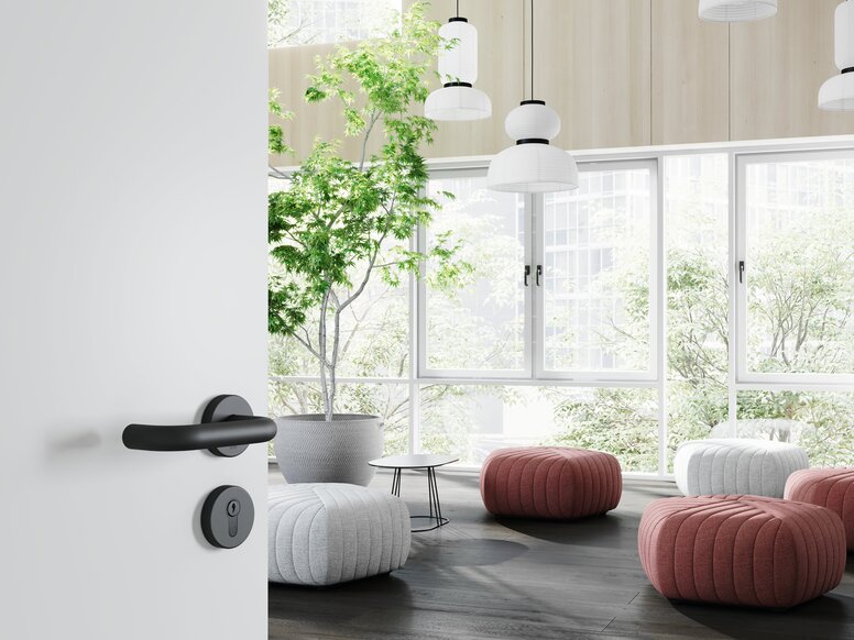 Black lever handle with a round rose on a white door, with large windows, plants and reddish seating in the bright lounge area beyond