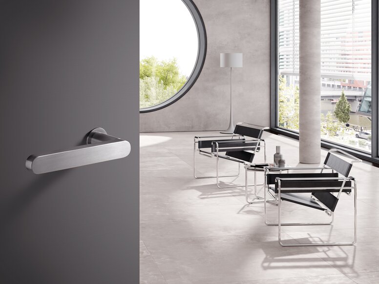HEWI Range 270 lever handles A stainless steel door handle on a grey door; behind it, a bright room with chairs