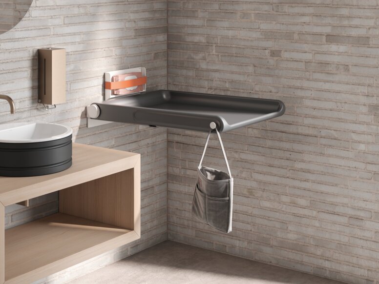 A hinged baby changing table with a side pocket for storage and a hook, mounted on a light-coloured brick wall