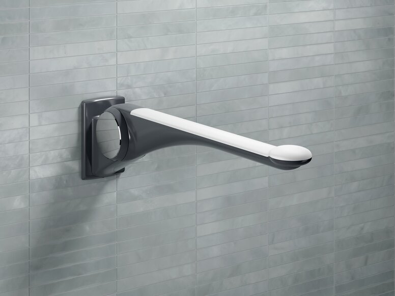 A hinged support rail made of dark grey synthetic material with an ergonomic white handle, fitted to a light grey tiled wall
