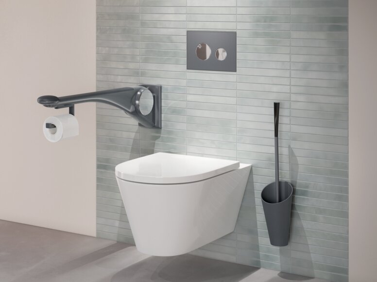 Wall-hung WC with a side support rail, an integrated toilet roll holder and a toilet brush mounted on a green tiled wall