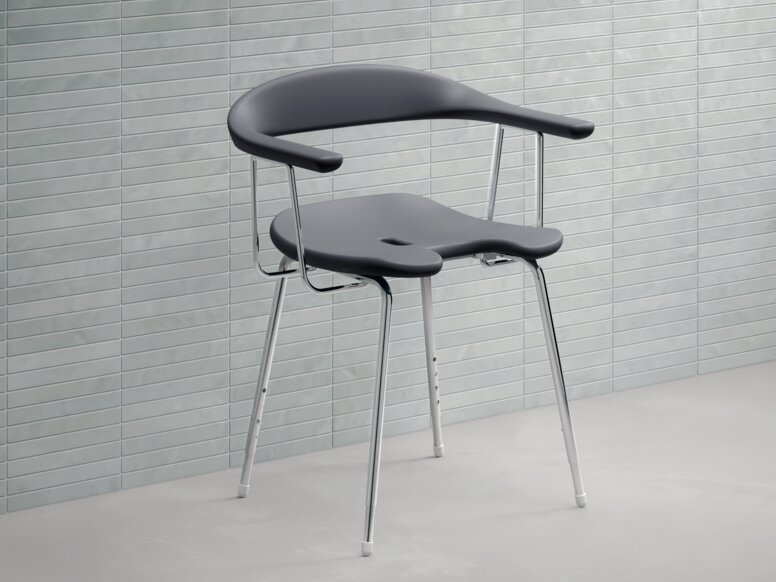Shower chair made of dark grey synthetic material with a semi-circular backrest, armrests and chrome-plated legs against a light grey tiled wall