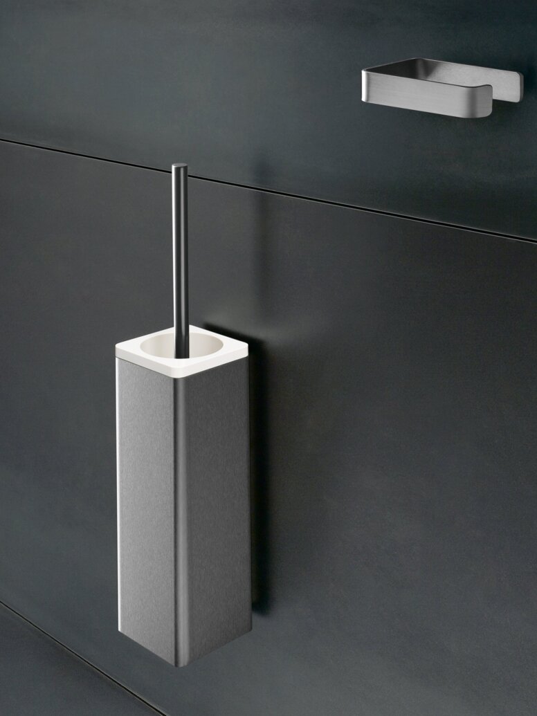 WC-Bürstengarnitur aus Edelstahl mit rechteckigem Behälter und Bürstengriff an einer dunkelgrauen Wand, darüber ein kleiner Wandhalter