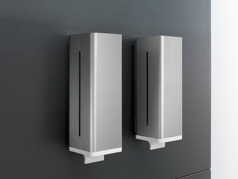 HEWI Series 805 Soap Dispensers Two rectangular soap dispensers with silver-grey fronts, narrow vertical viewing windows and white dispensing outlets, mounted side by side on a dark grey wall